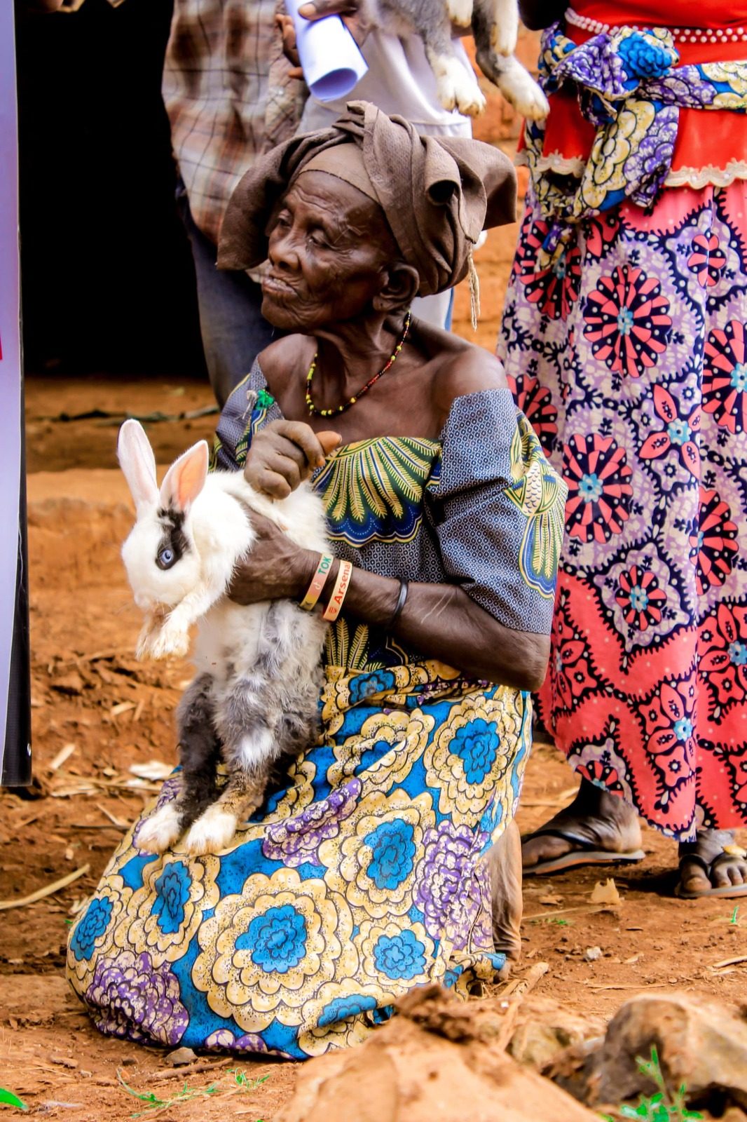 A woman seated on red soil holding a white and grey rabbit