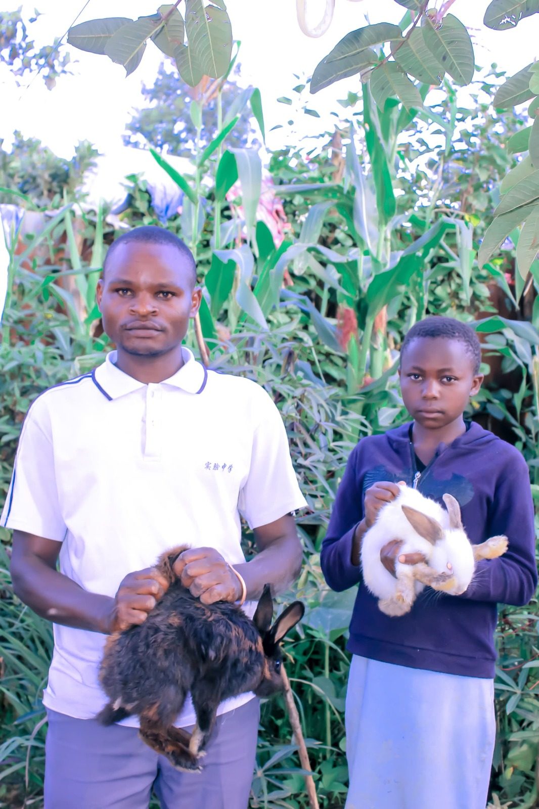 A man and a boy each holding a rabbit in front of tall crops